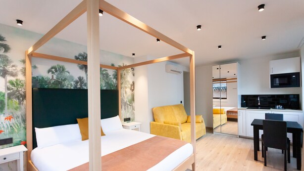 Una habitación de hotel moderna con una cama con dosel, un sillón amarillo, una mesa de comedor con sillas y una zona de cocina.