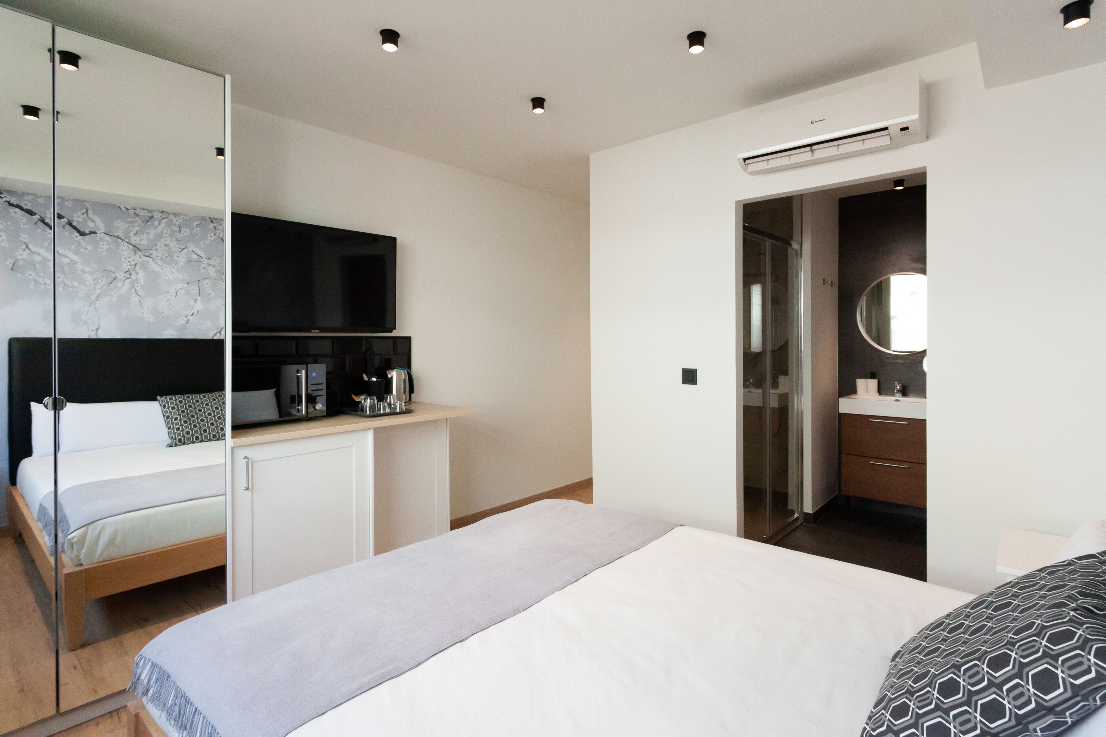A modern hotel room with a bed, a desk with a coffee maker, a TV, and a bathroom with a mirror and sink.