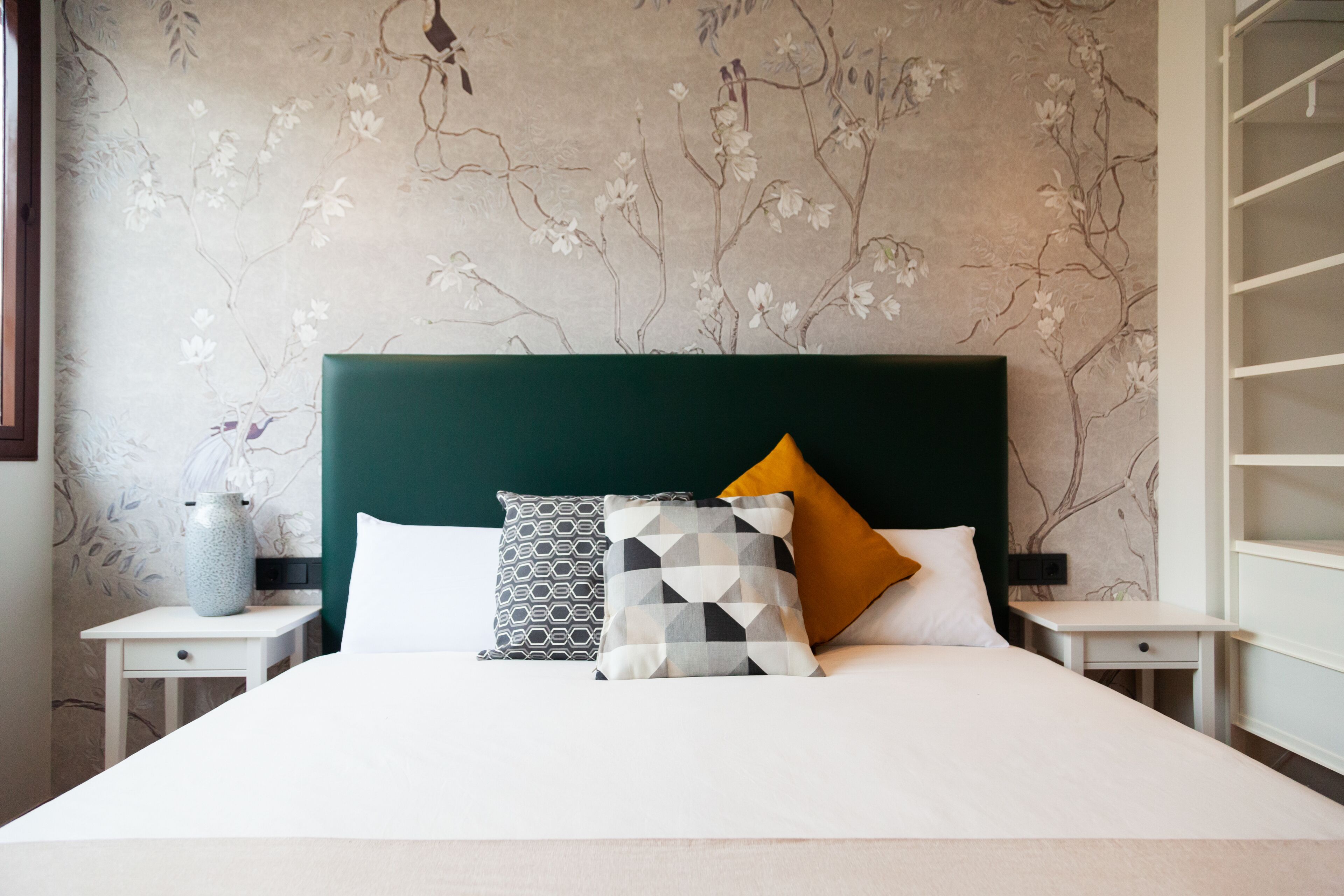A neatly made bed with a geometric-patterned pillow, a vase, and a wall with a floral design.