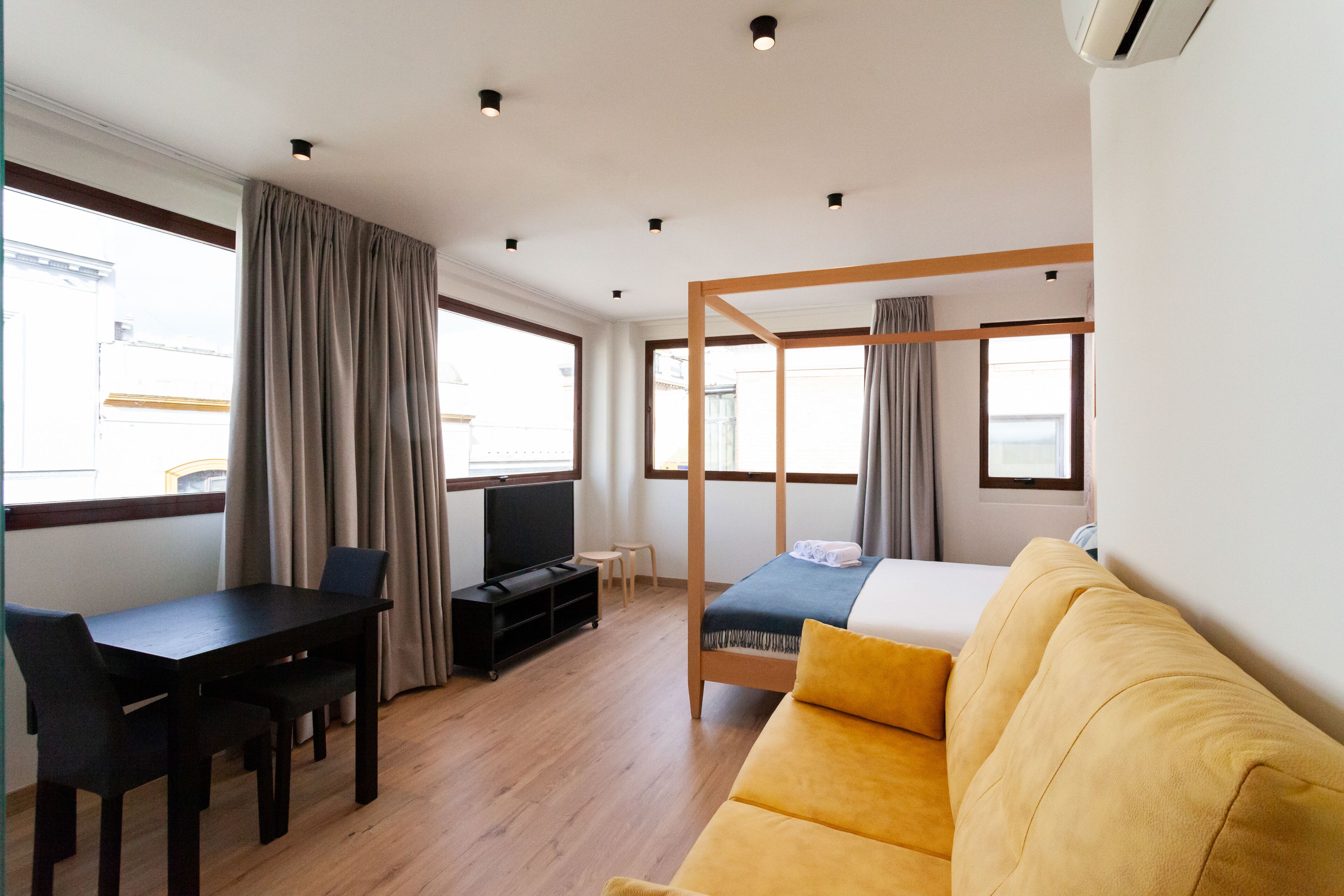 Una habitación de hotel moderna con piso de madera, un sofá amarillo, un televisor, una mesita y un ventanal grande con cortinas.