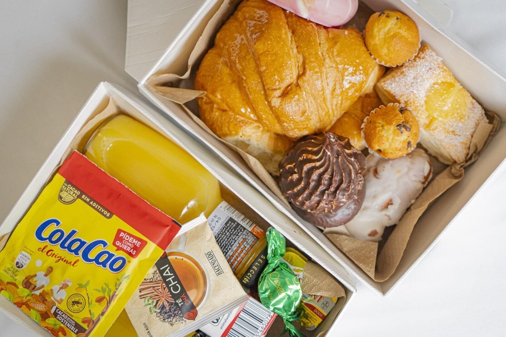 Two open boxes containing various food items including a chocolate-covered pastry, a croissant, and a yellow beverage.