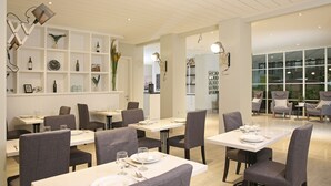 A modern dining area with white tables and grey chairs, a white shelving unit with decorative items, and a view through large windows. - Havenwood Residence TB Simatupang (Jakarta)
