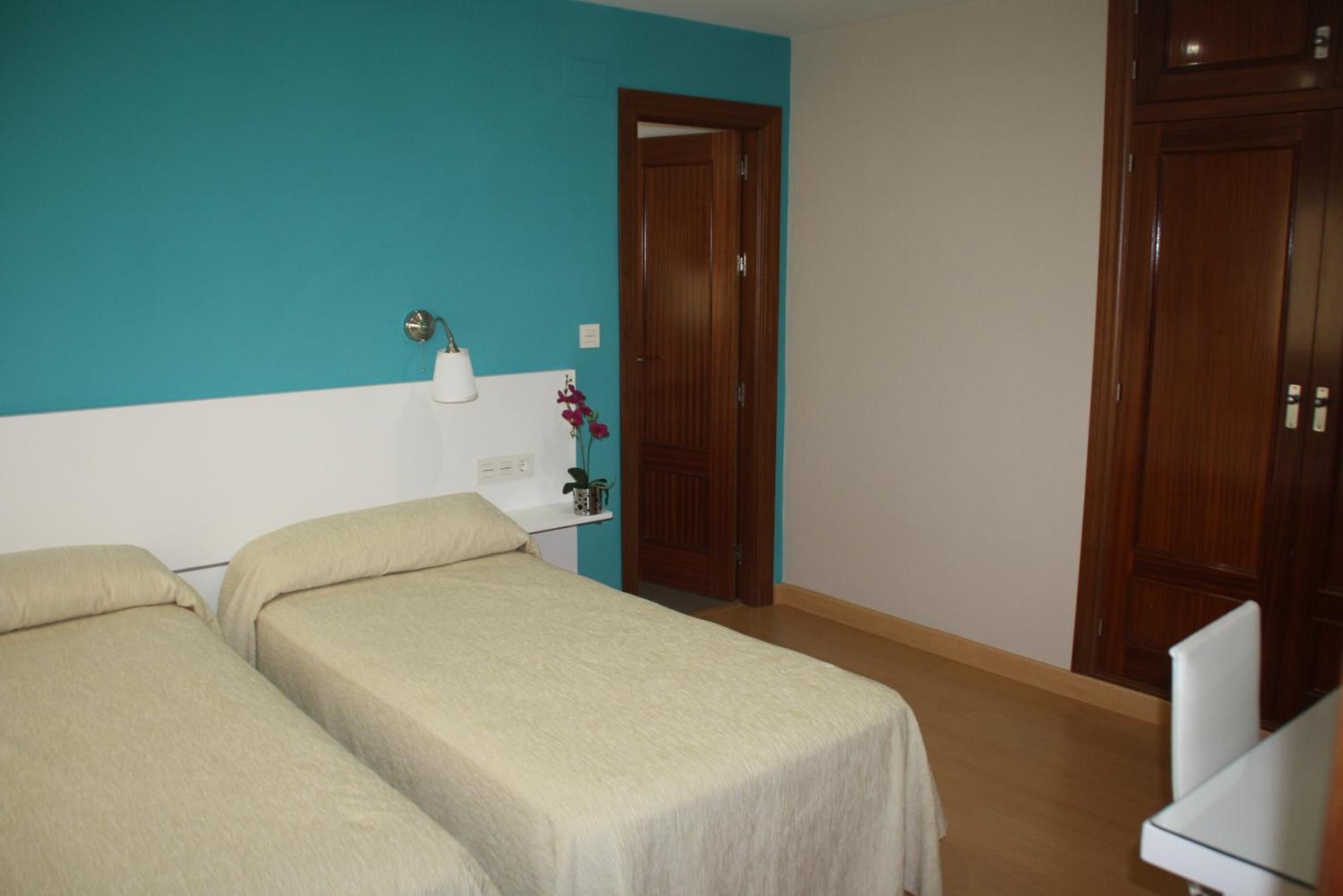 A room with two beds, a teal wall, a wooden door, and a small shelf with a plant.