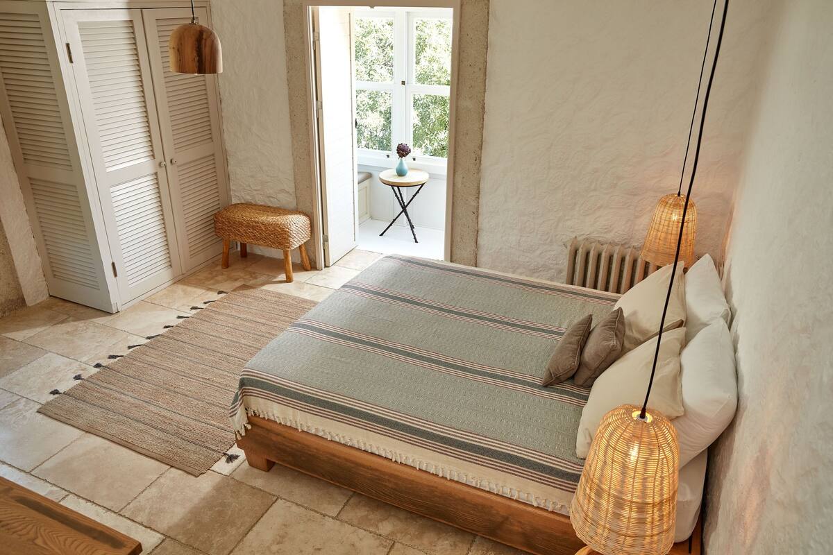 Ein Schlafzimmer mit einem Bett, einem kleinen runden Tisch, einer Korbsitzbank und Blick ins Freie durch ein Fenster.