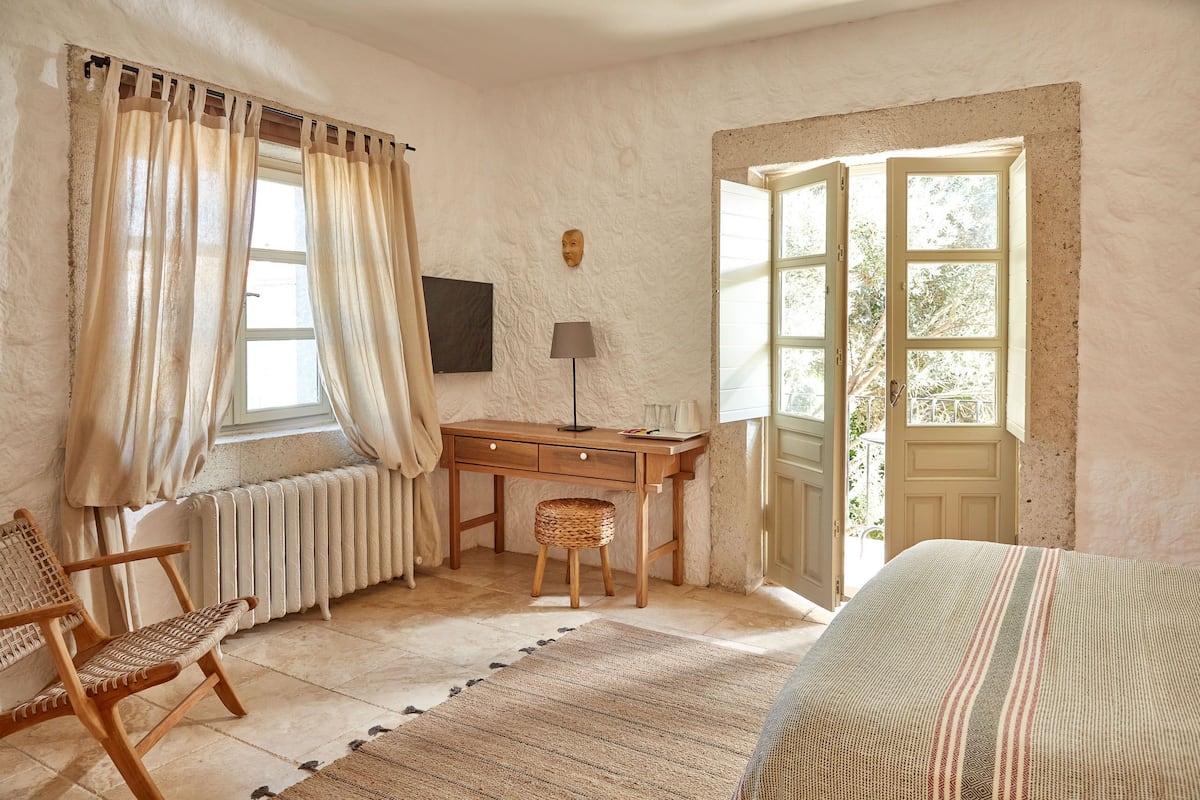 Ein Schlafzimmer mit Holzschreibtisch, einem Sessel, einer Heizung, einem Fenster mit Vorhängen, einer Lampe und einer Tür, die nach draußen führt.
