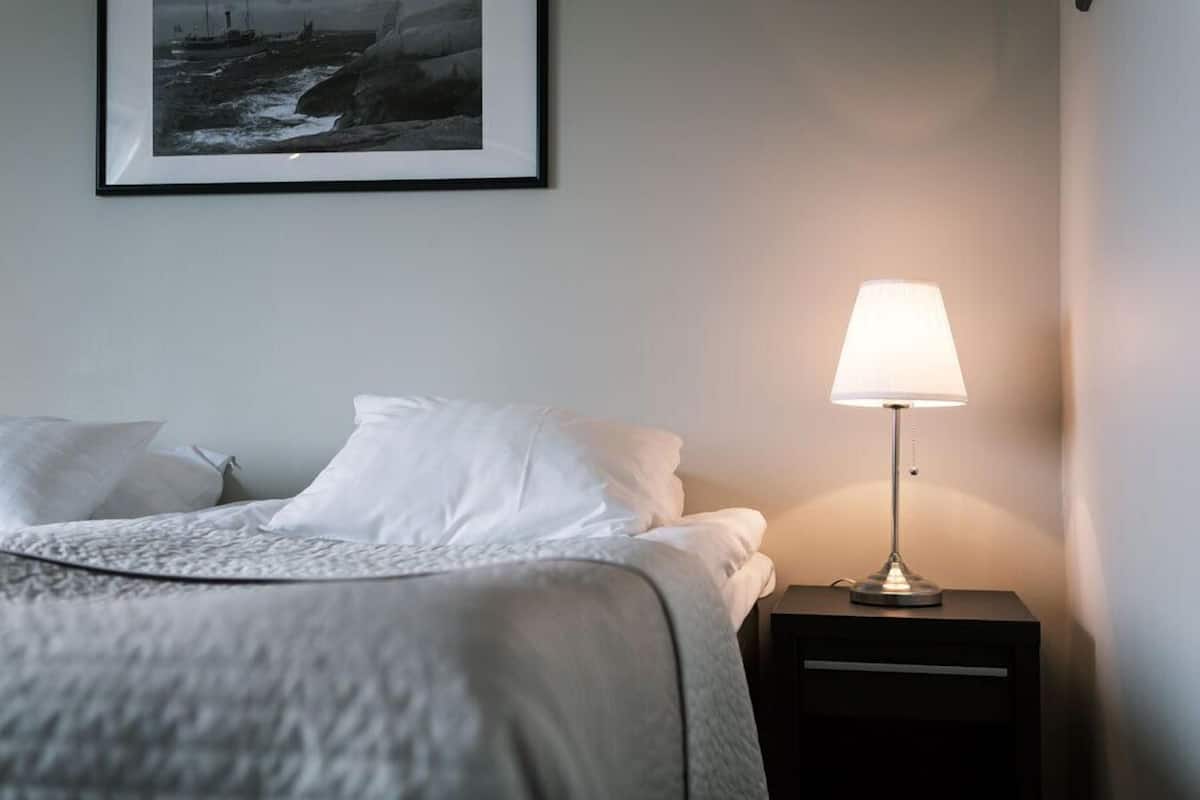A neatly made bed with white linens, a framed picture above it, and a bedside table with a lamp.