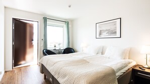 Una habitación de hotel con cama, escritorio, silla, ventana con cortinas, un cuadro en la pared y mesita de noche con lámpara.