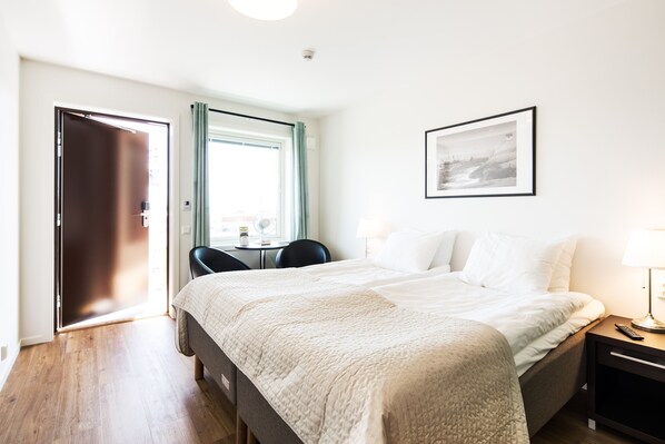 Une chambre d’hôtel comprenant un lit, un bureau, une chaise, une fenêtre avec des rideaux, un tableau encadré au mur et une table de chevet avec une lampe.