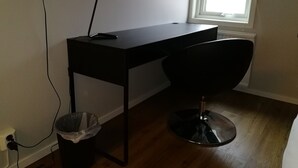 Un pequeño escritorio oscuro con una silla, un cubo de basura y una ventana con cortinas blancas.