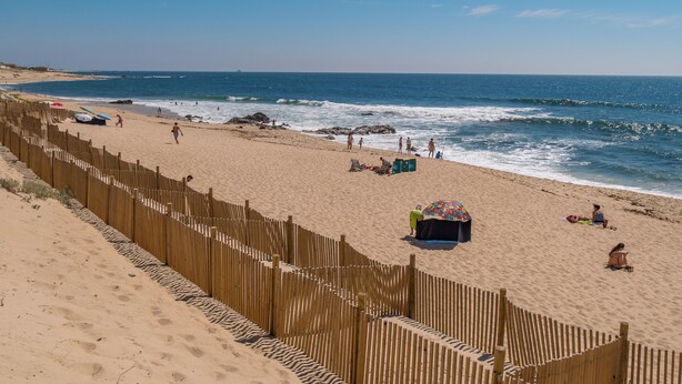 Una playa con una valla de madera, gente caminando y sillas de playa.