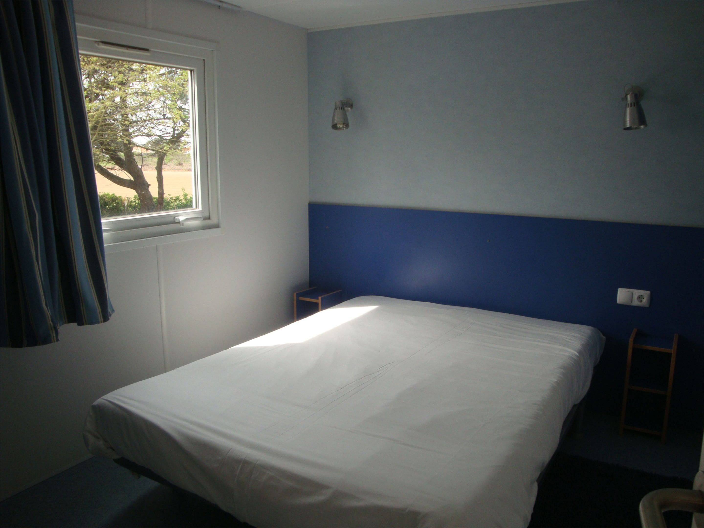 A bedroom with a single bed, a window with curtains, a small table, and a wall-mounted light fixture.