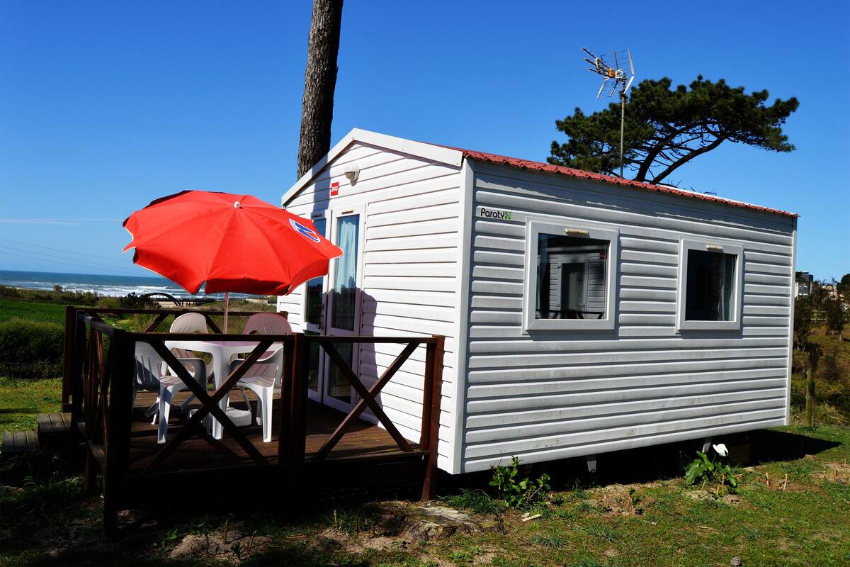 A small white mobile home with a red roof, a deck with chairs, and a red umbrella.