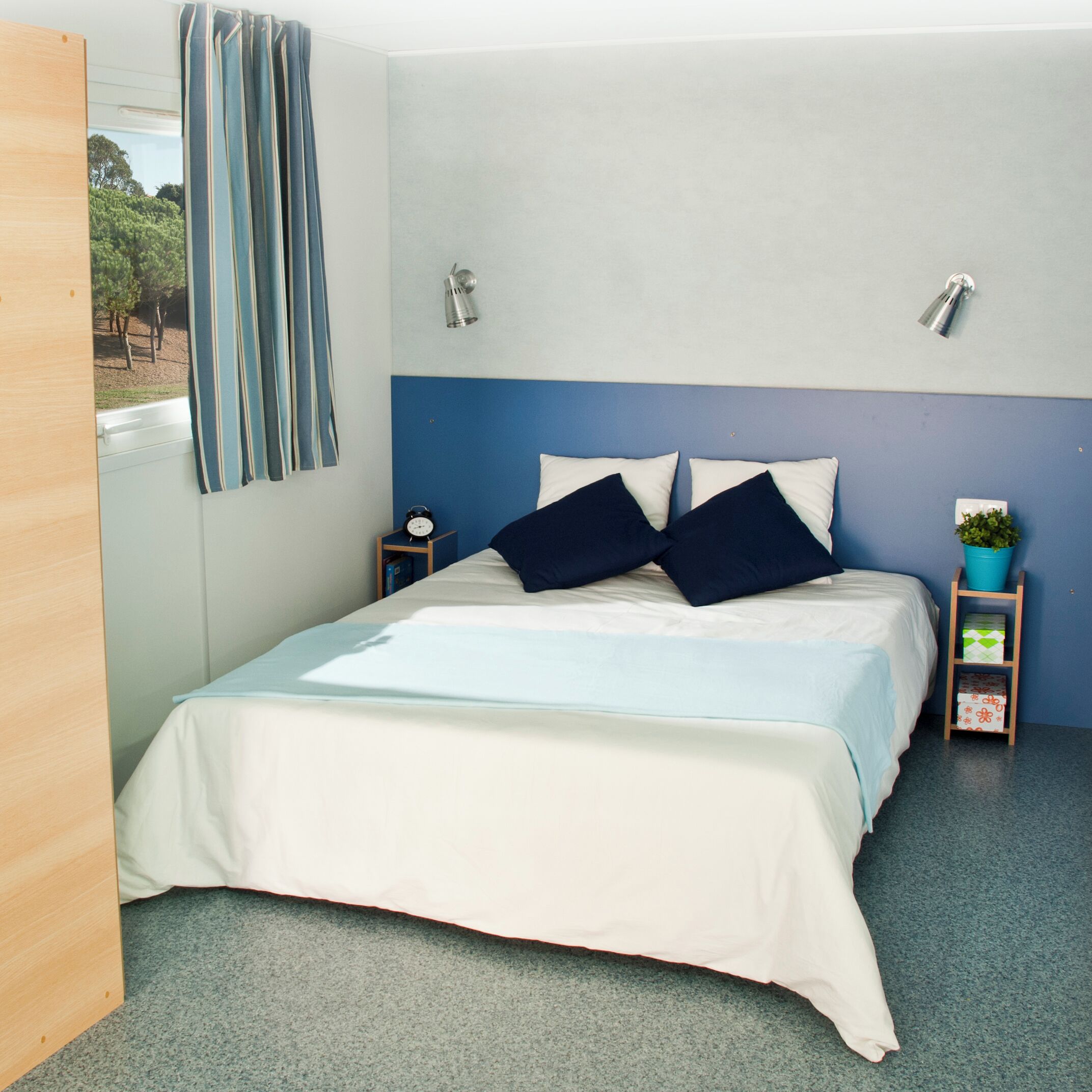 A bedroom with a bed, a nightstand, a wall-mounted lamp, a window with curtains, and a small plant on a side table.