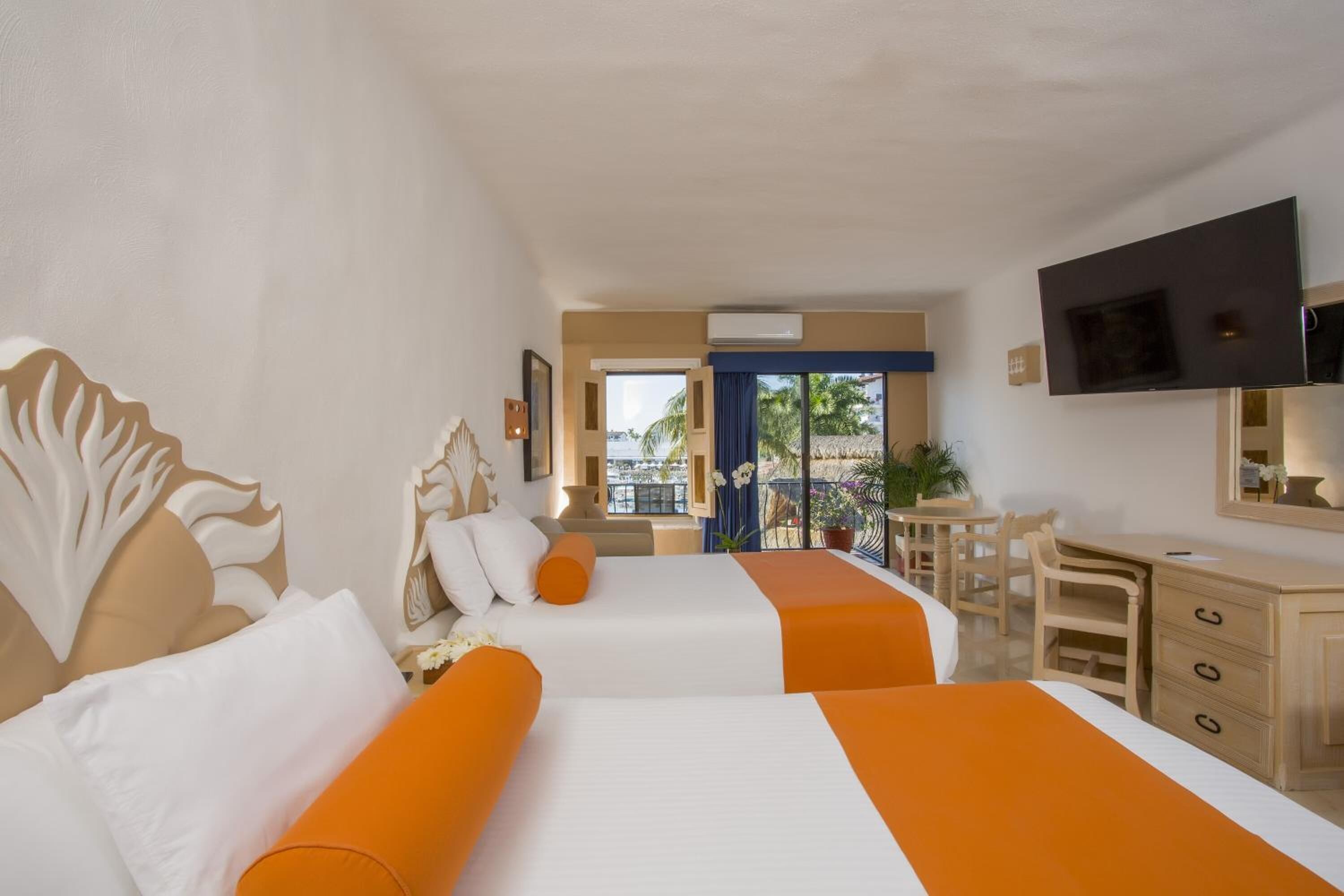 Habitación de hotel con dos camas, un escritorio, una silla, un televisor y un balcón con vistas a palmeras.