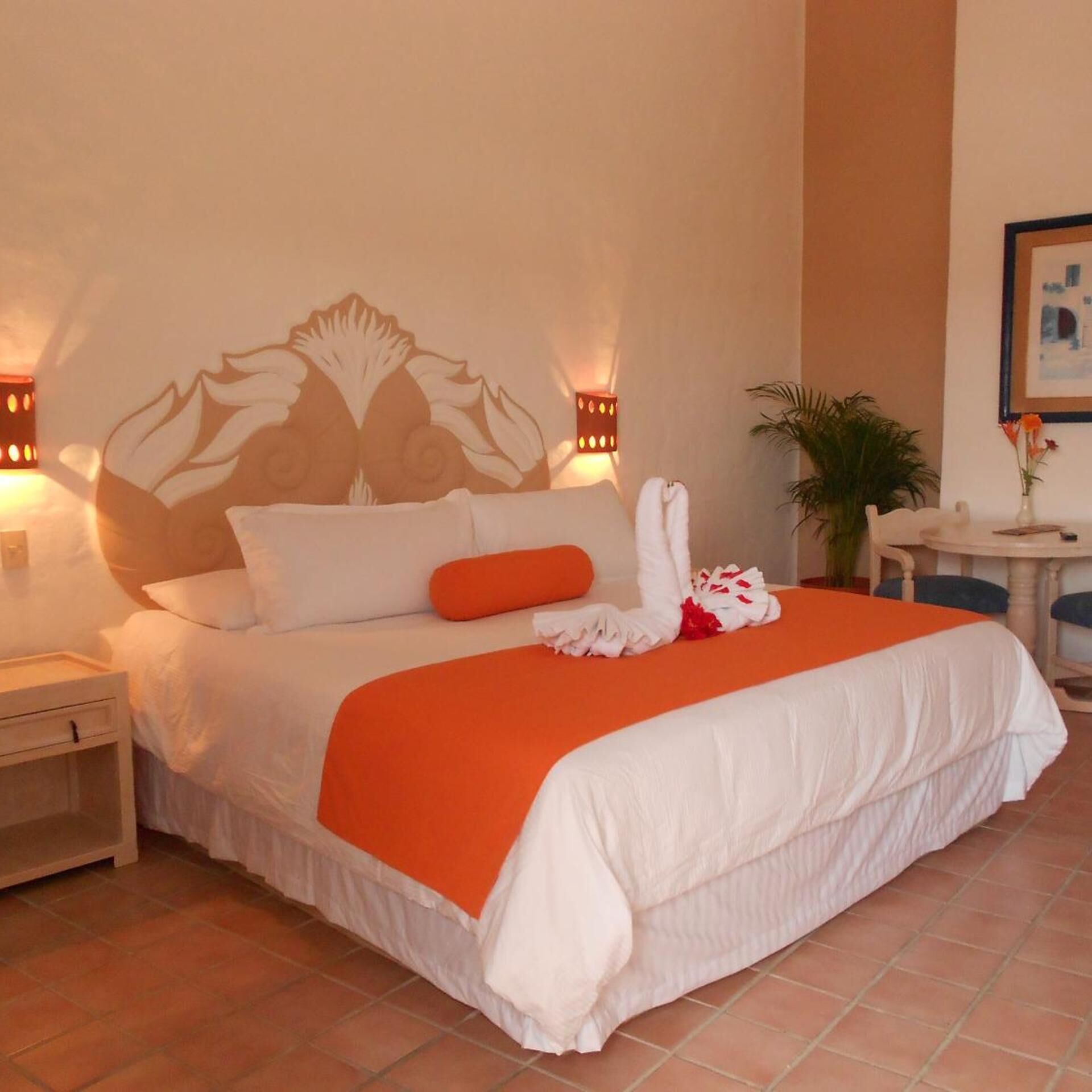 A hotel room with a bed, a nightstand, a wall-mounted lamp, a small table with a vase, and a potted plant.