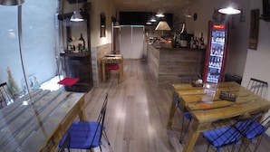 L’intérieur d’un restaurant présente des tables en bois et des chaises rembourrées de tissu bleu, un coin bar avec des bouteilles exposées, et un réfrigérateur rempli de boissons.