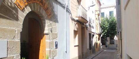A narrow street with a building labeled "HOSTAL" featuring a stone facade, a wooden door, and a small potted plant on the sidewalk.
