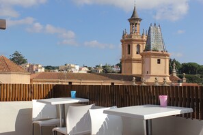 Une terrasse avec des tables et des chaises blanches, une clôture en bois et une vue sur un bâtiment historique doté d’une tour.
