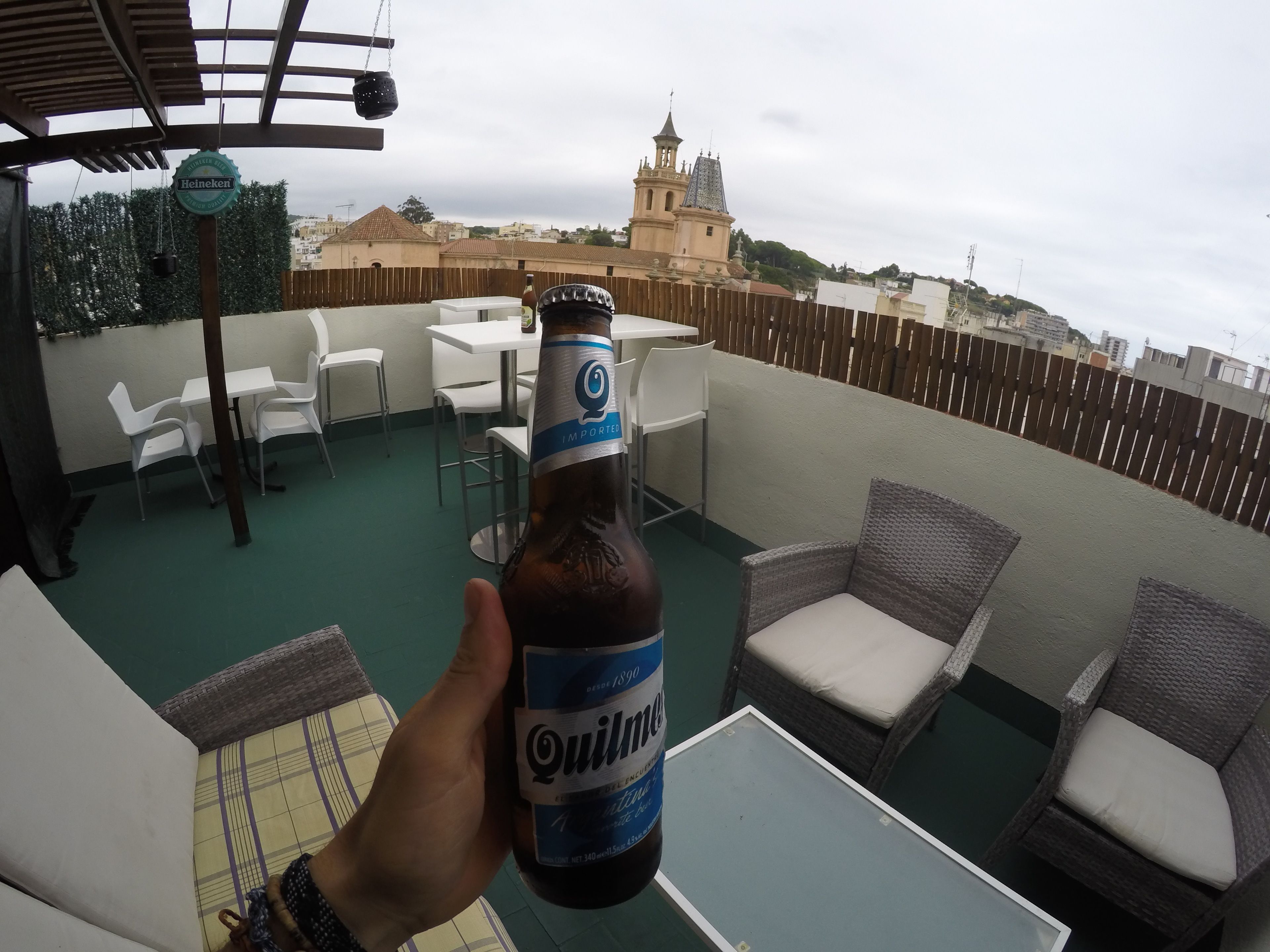 Una botella de cerveza Quilmes en una terraza en la azotea con sillas y mesas blancas, una valla de madera y vistas a una iglesia con un campanario.