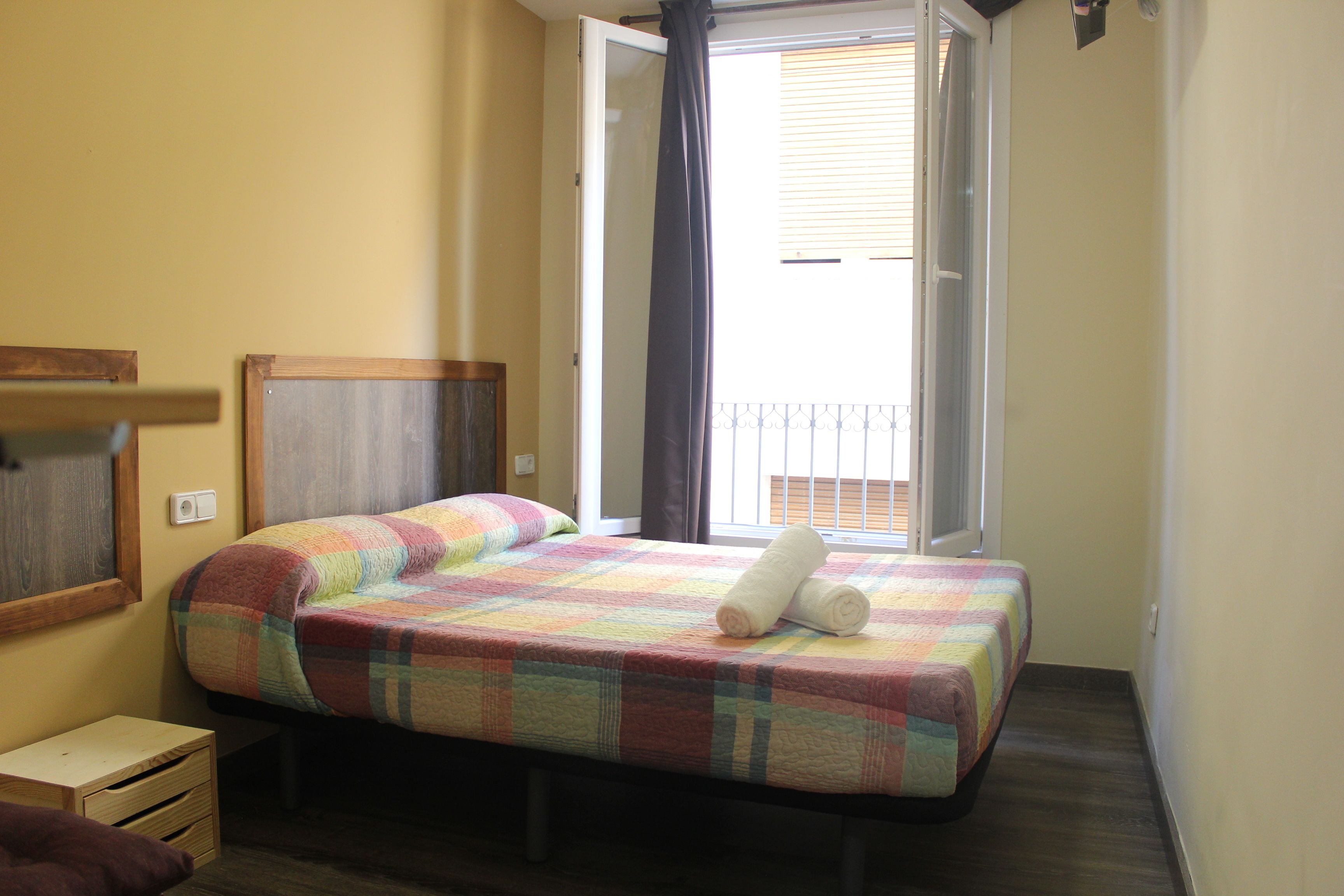 A bedroom with a bed, a small wooden nightstand, a window with curtains, and a view of the outside balcony.