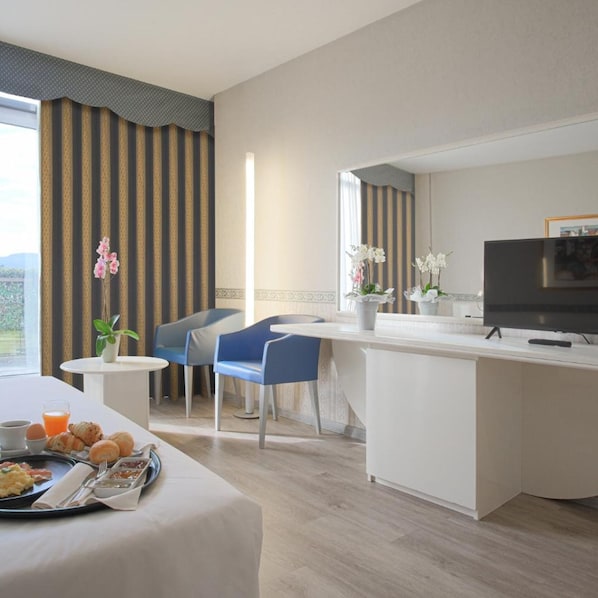 Ein Hotelzimmer mit Bett, Frühstückswagen, zwei Stühlen, einem kleinen Tisch mit Pflanze, einem Fernseher und einem großen Fenster mit Vorhängen.