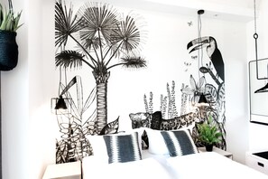 Un dormitorio con cama, almohadas, una planta colgante y un mural en la pared.