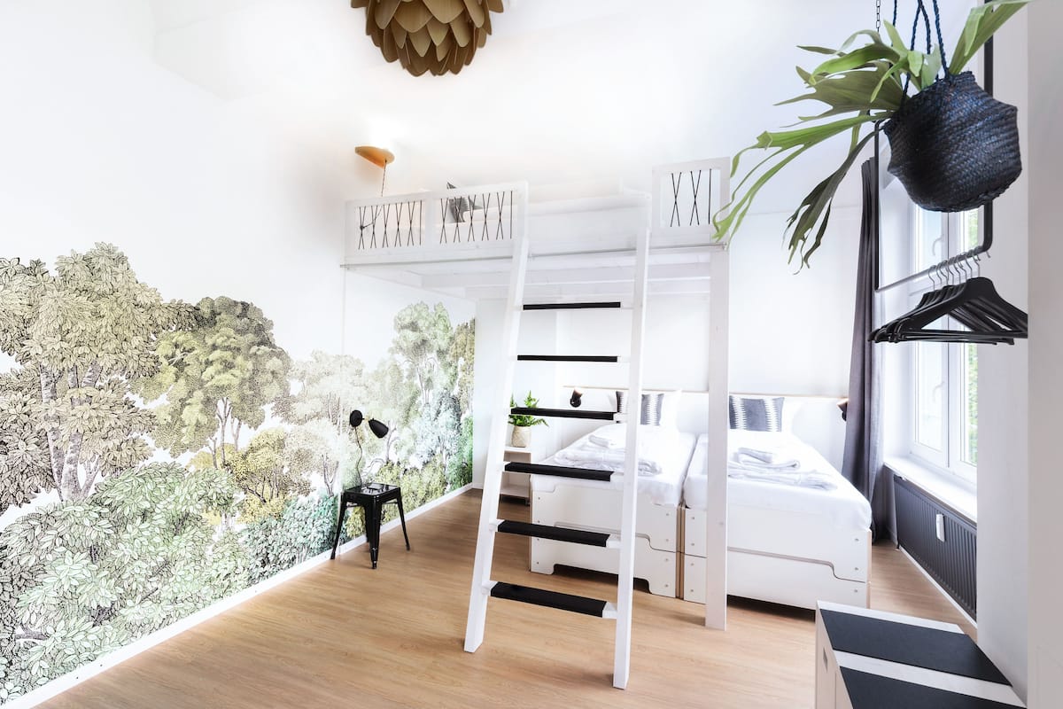 A modern bedroom with a bunk bed, a ladder, a hanging plant, and a view of a forest mural.