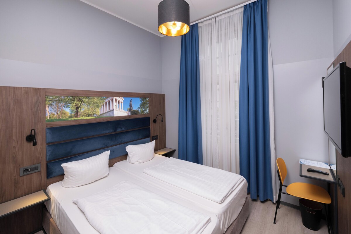 A hotel room with a bed, a desk, a chair, a TV, and a window with curtains.