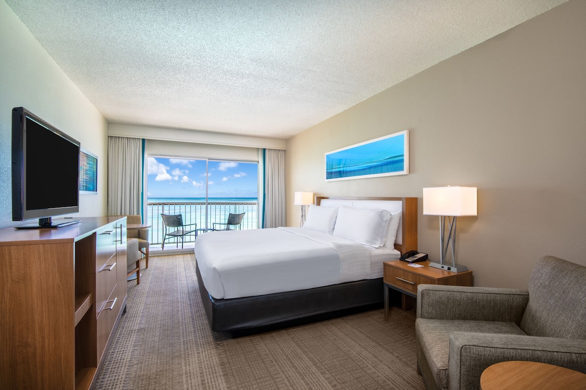 A hotel room with a large bed, a TV, a desk, a chair, and a balcony with a view of the sky.