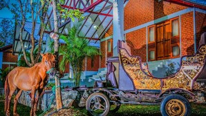 Un cheval se tient à côté d’une calèche ornée de motifs dorés complexes, garée devant un bâtiment en briques doté de grandes fenêtres et d’une structure de toit en verre.