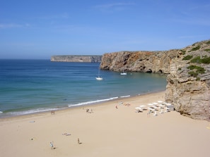 Ein Strand mit blauem Himmel, ruhigem Wasser und felsigen Klippen.