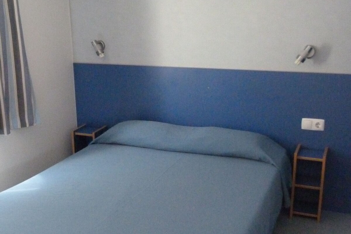Ein Zimmer mit einem Bett, einem kleinen Tisch, einem Stuhl und einer Wandleuchte.