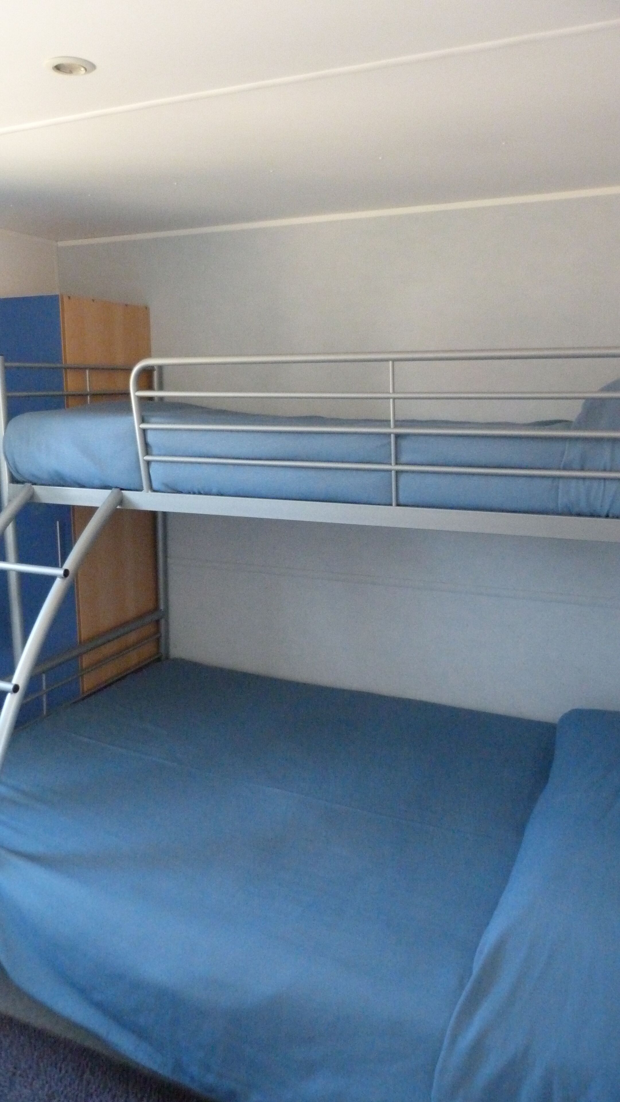 Un lit superposé avec un matelas bleu, des barreaux en métal et un panneau en bois sur le côté gauche.