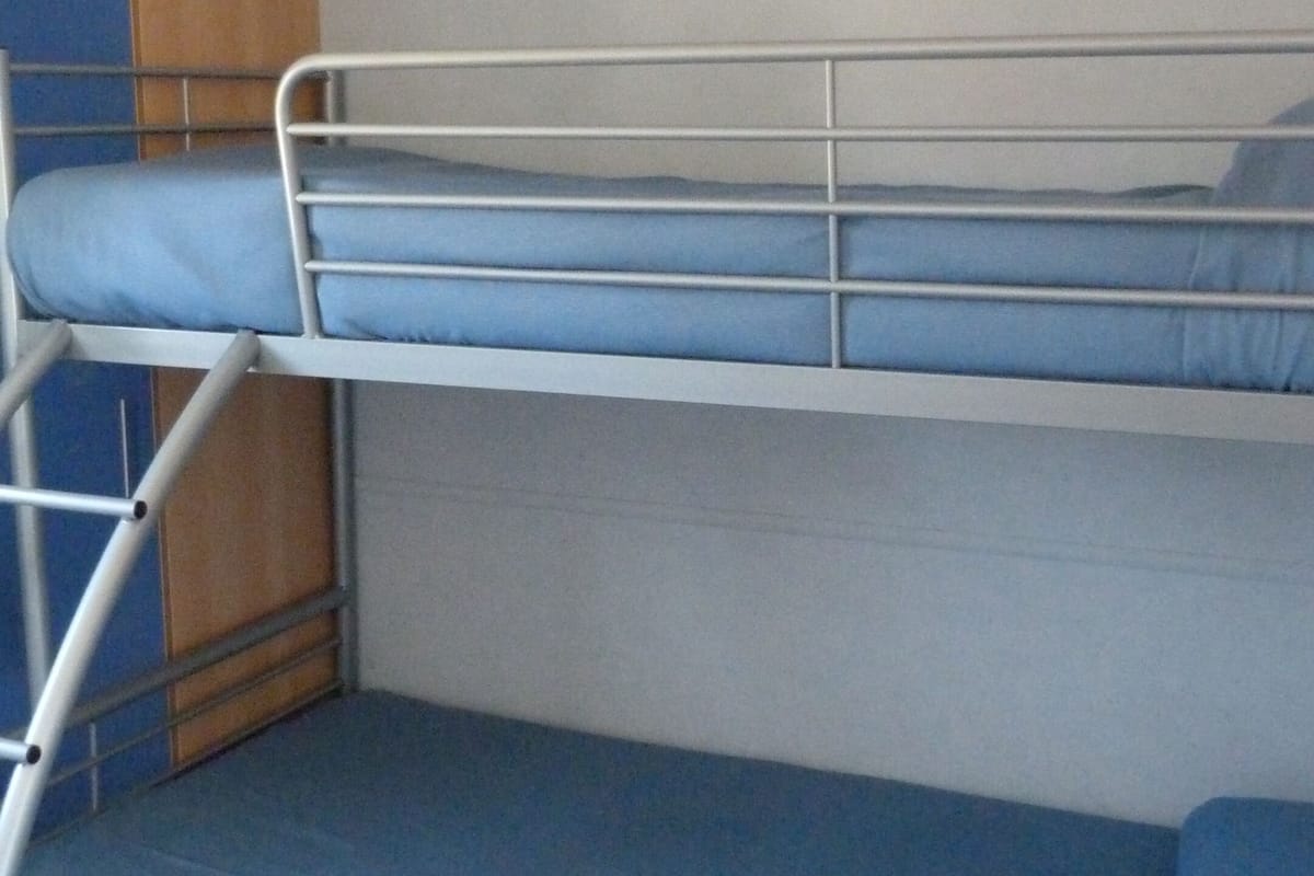 Una cama litera con colchón azul, barandillas de metal y un panel de madera a la izquierda.