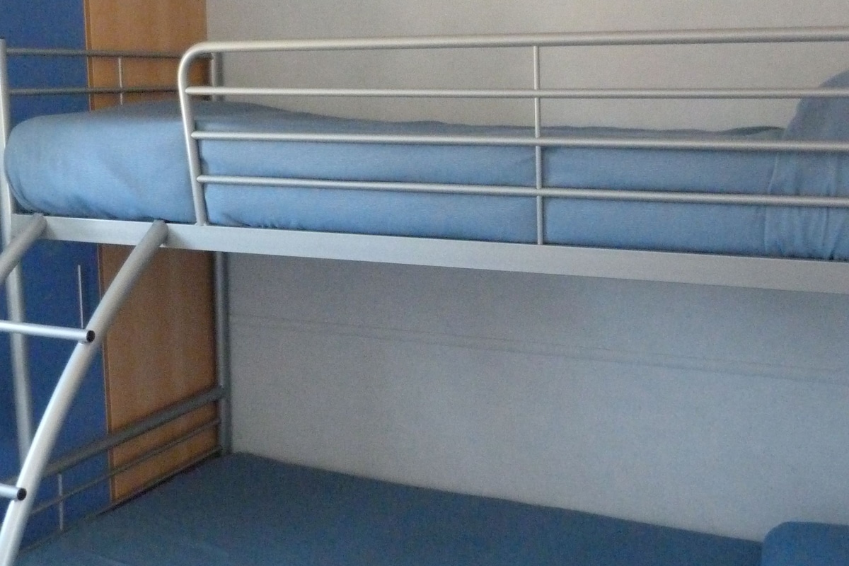 Ein Stockbett mit blauer Matratze, Metallgitter und einer Holzplatte links.