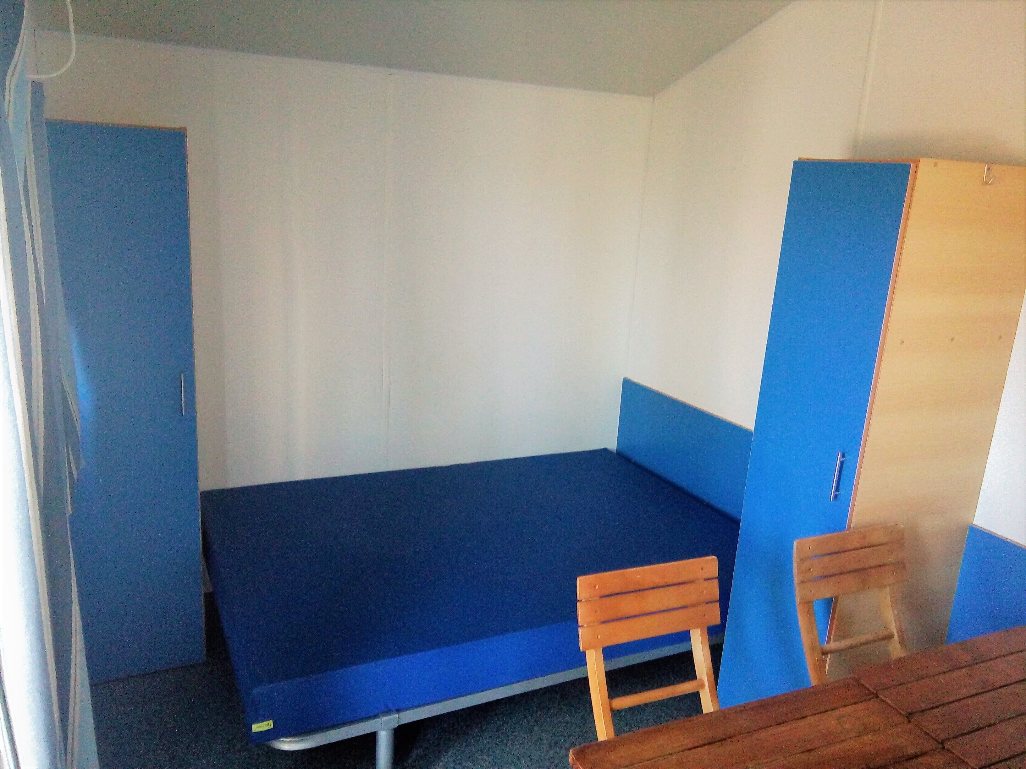 Une petite chambre avec un lit simple, un matelas bleu, deux chaises en bois et une porte en bois.
