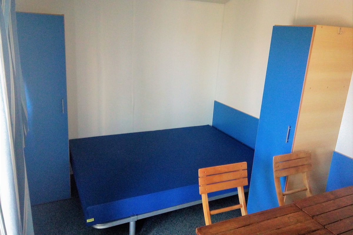 Una habitación pequeña con una cama individual, colchón azul, dos sillas de madera y una puerta de madera.