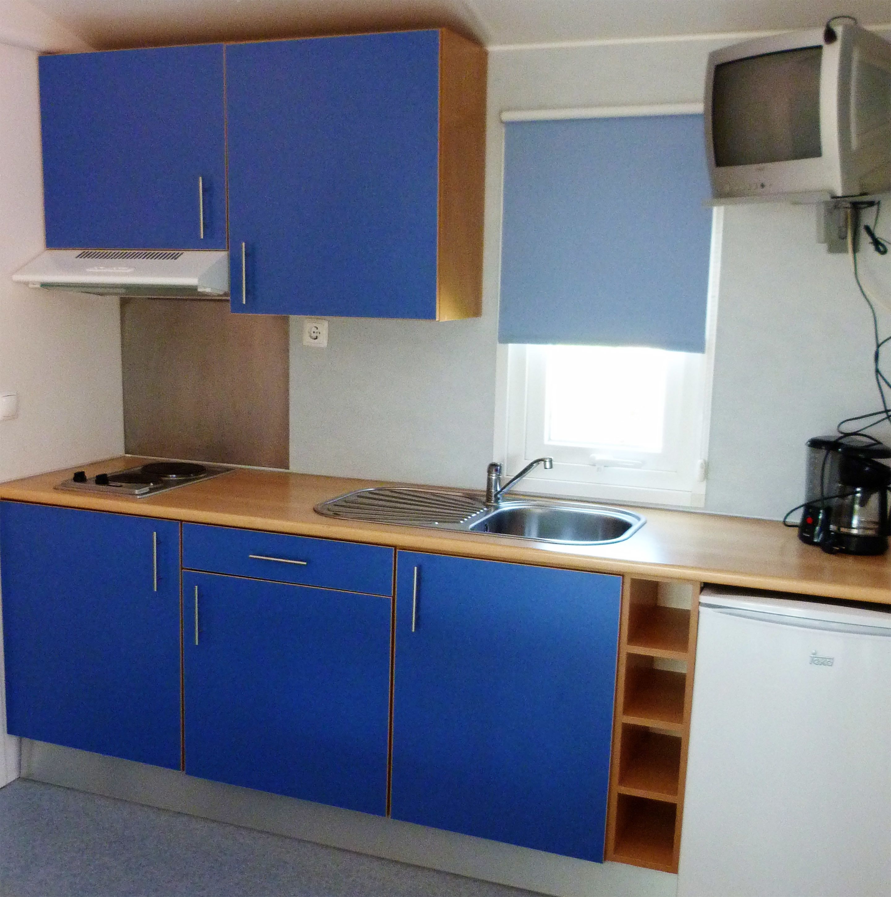 Una kitchenette compacta con gabinetes azules, un lavamanos pequeño, una estufa y un televisor montado en la pared.