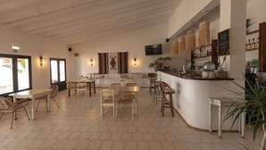Un vaste espace dédié à la salle à manger, doté d’un sol carrelé à motifs hexagonaux, d’une table blanche centrale entourée de chaises en osier, et d’un coin bar avec un comptoir et des luminaires suspendus.