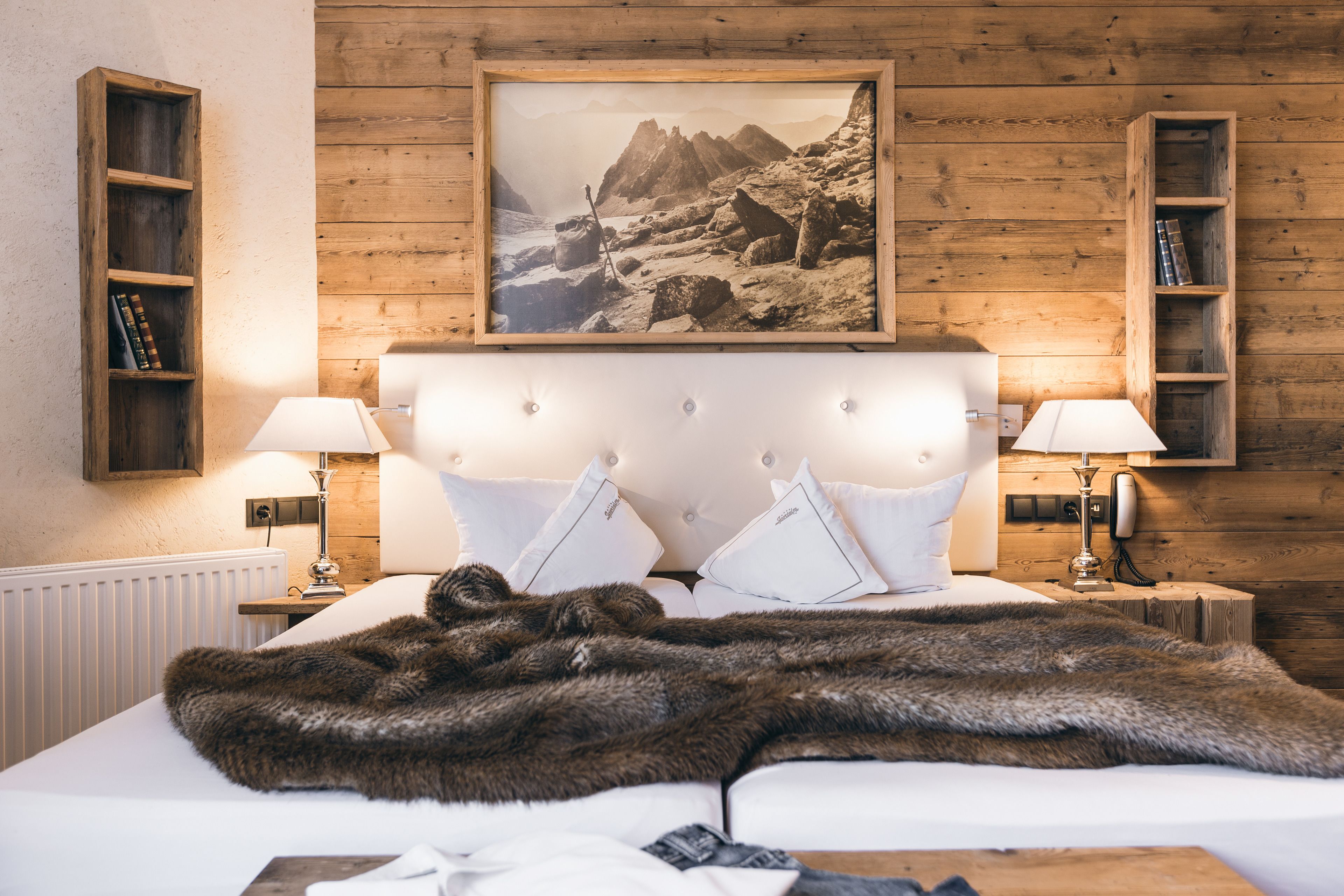 A bedroom with a wooden headboard, white bedding, two bedside lamps, a framed picture of a mountainous landscape, and wooden shelves with books.