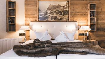 A bedroom with a wooden headboard, white bedding, two bedside lamps, a framed picture of a mountainous landscape, and wooden shelves with books.