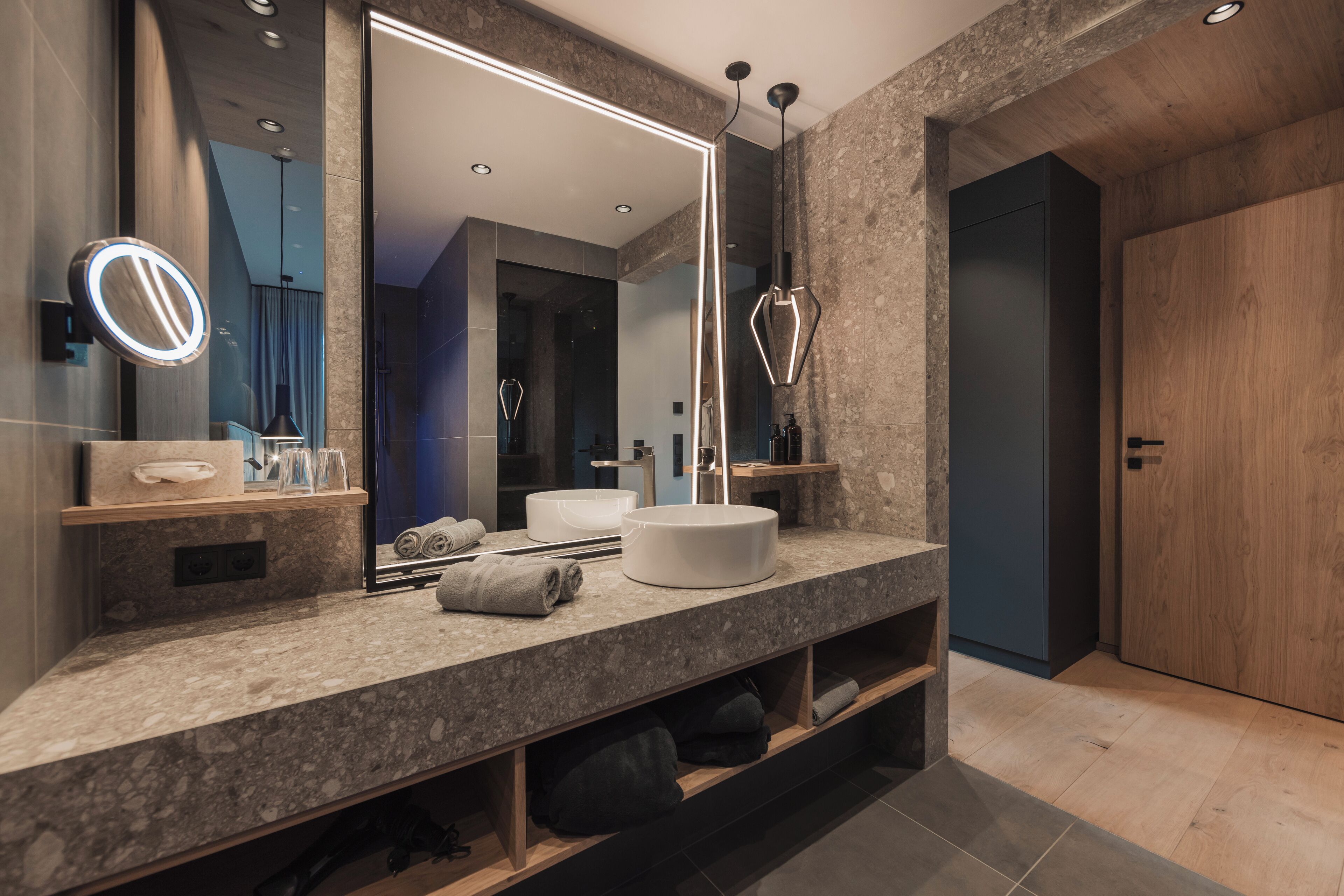 A modern bathroom with a large mirror, a round light, a white sink, and a wooden door.