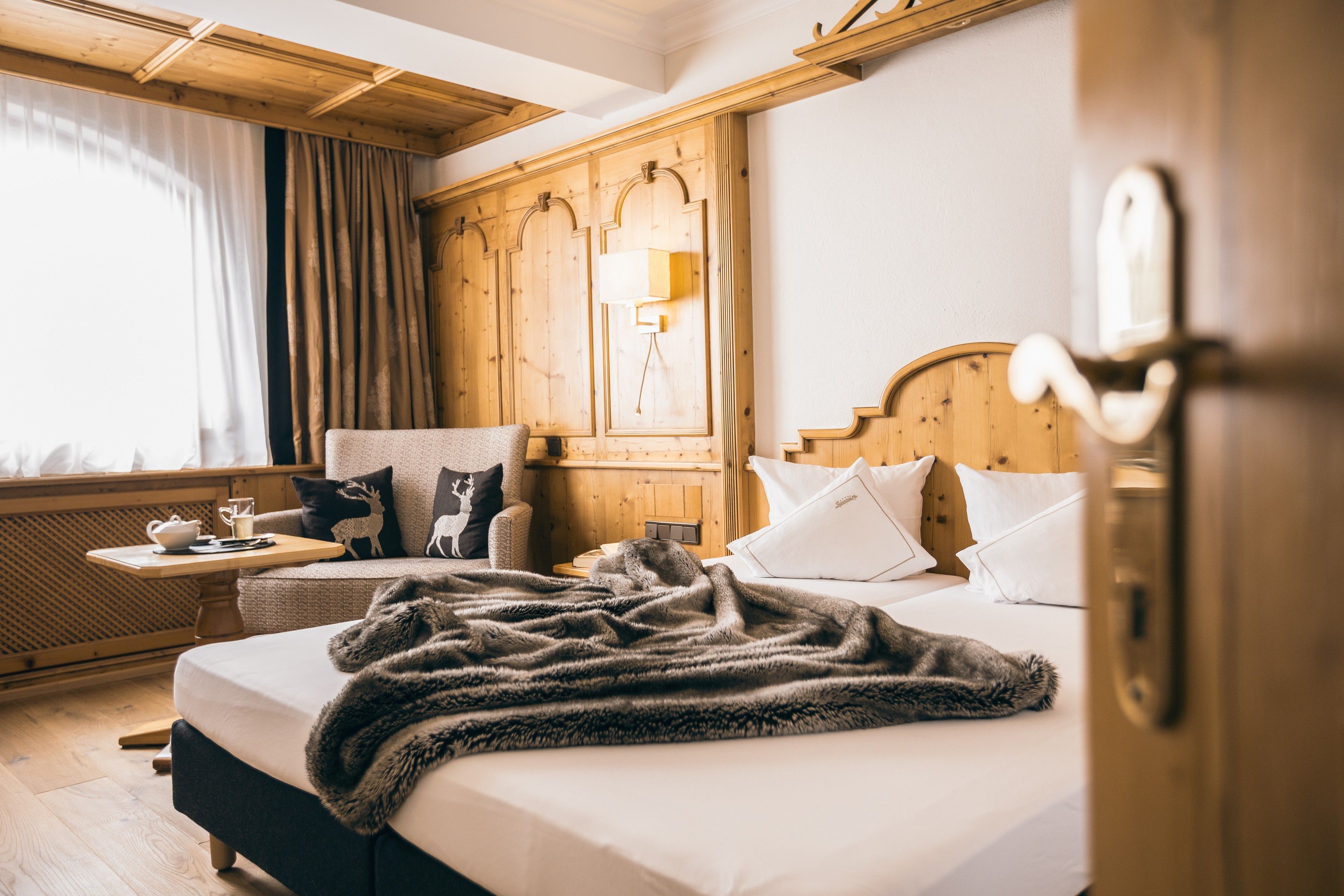 Ett mysigt hotellrum med en sänggavel i trä, en säng med ett mjukt täcke, ett litet bord med en kopp och ett fat samt ett fönster med gardiner.