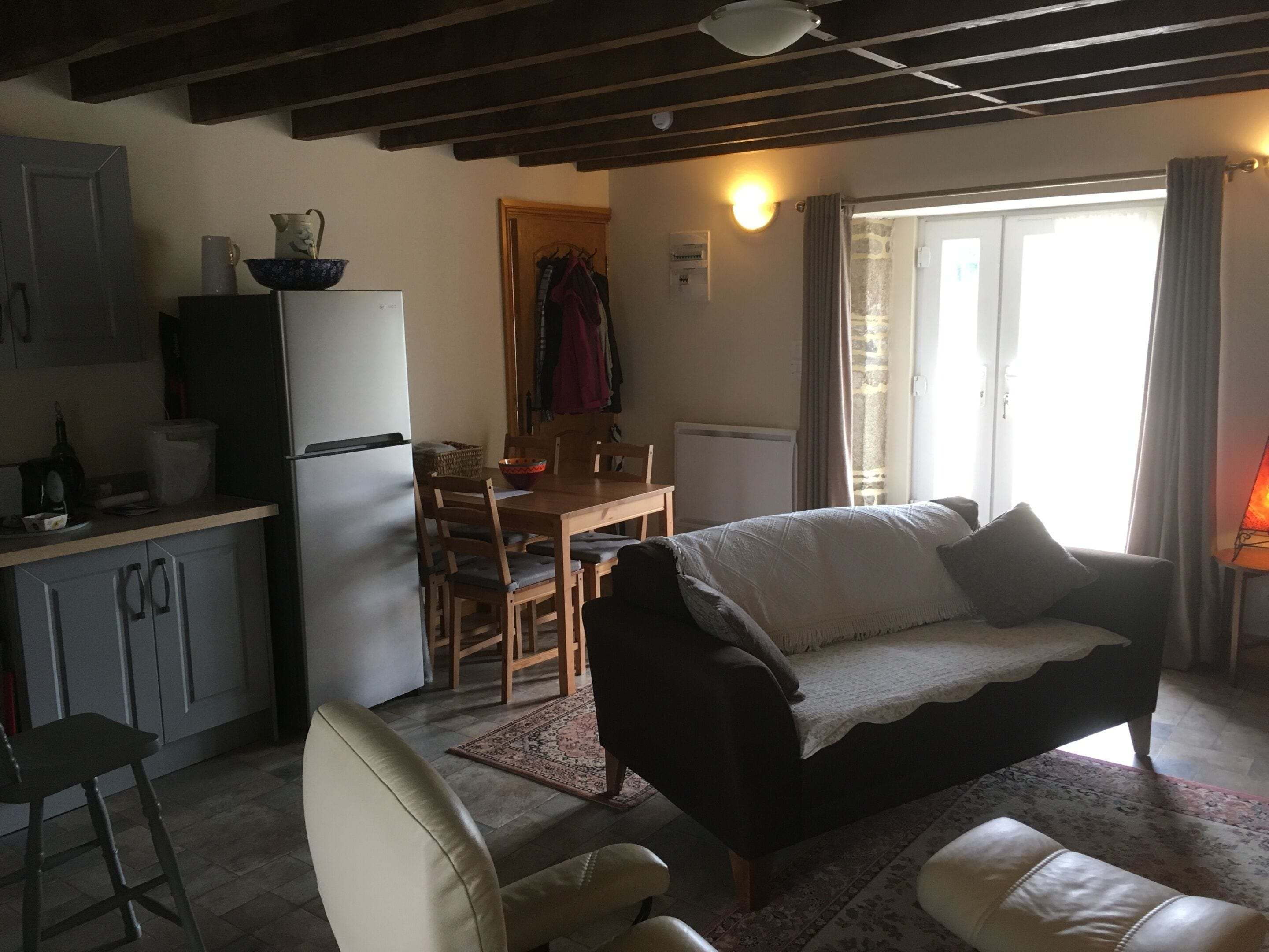 Un salon confortable avec un canapé, une table à manger avec des chaises, un coin cuisine avec des placards et un réfrigérateur, et une grande fenêtre avec des rideaux.