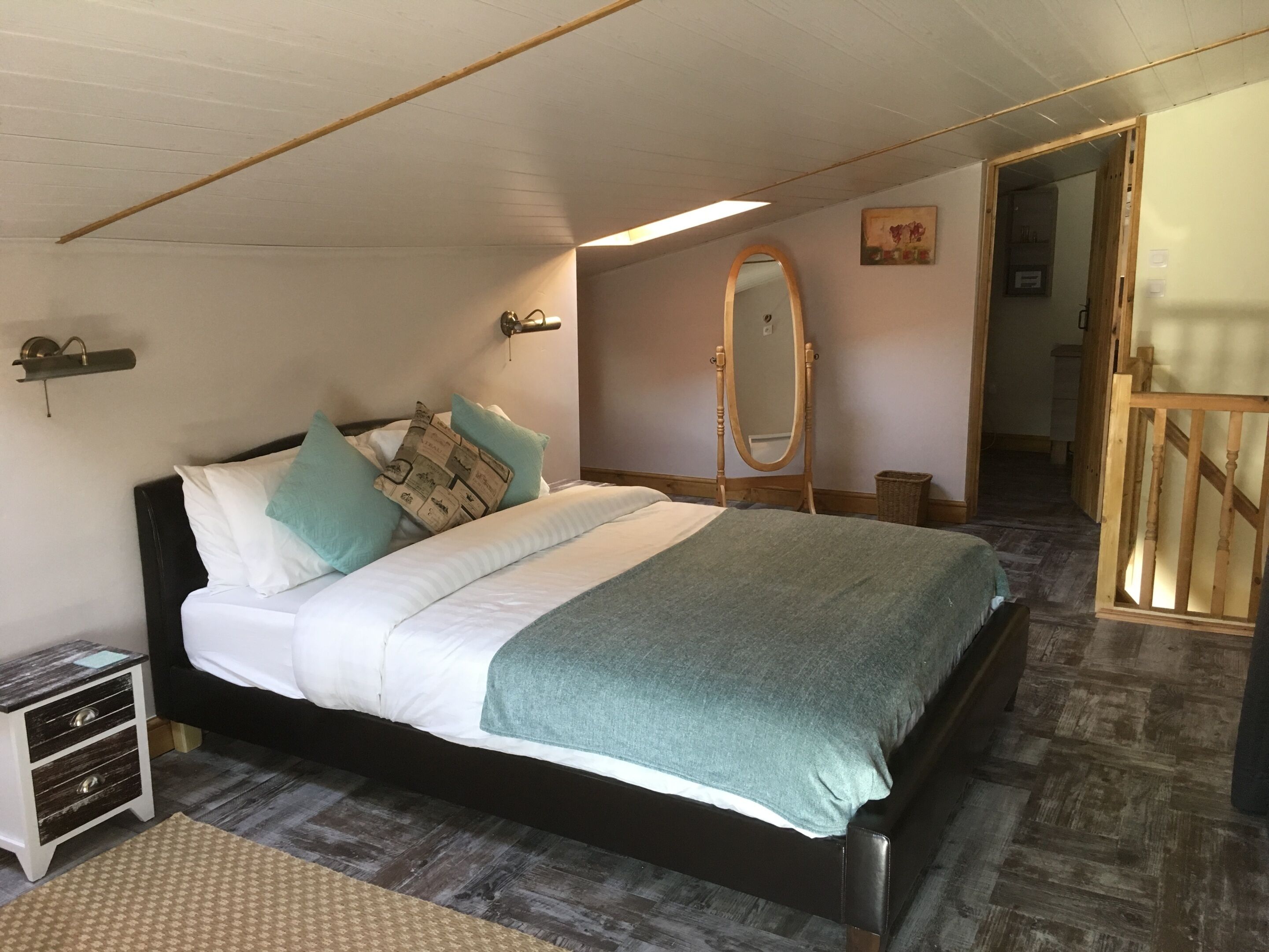 A bedroom with a bed, a wooden headboard, a bedside table, a mirror, and a wooden floor.