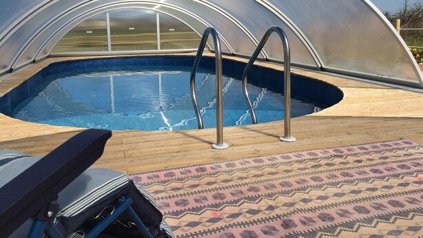 A swimming pool with a transparent roof, surrounded by a wooden deck and a patterned rug. - Le Bois Gautier (Savigny-le-Vieux)