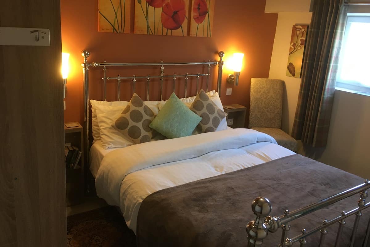 A neatly made bed with a metal headboard, a bedside table with a lamp, and a chair with a patterned cushion.