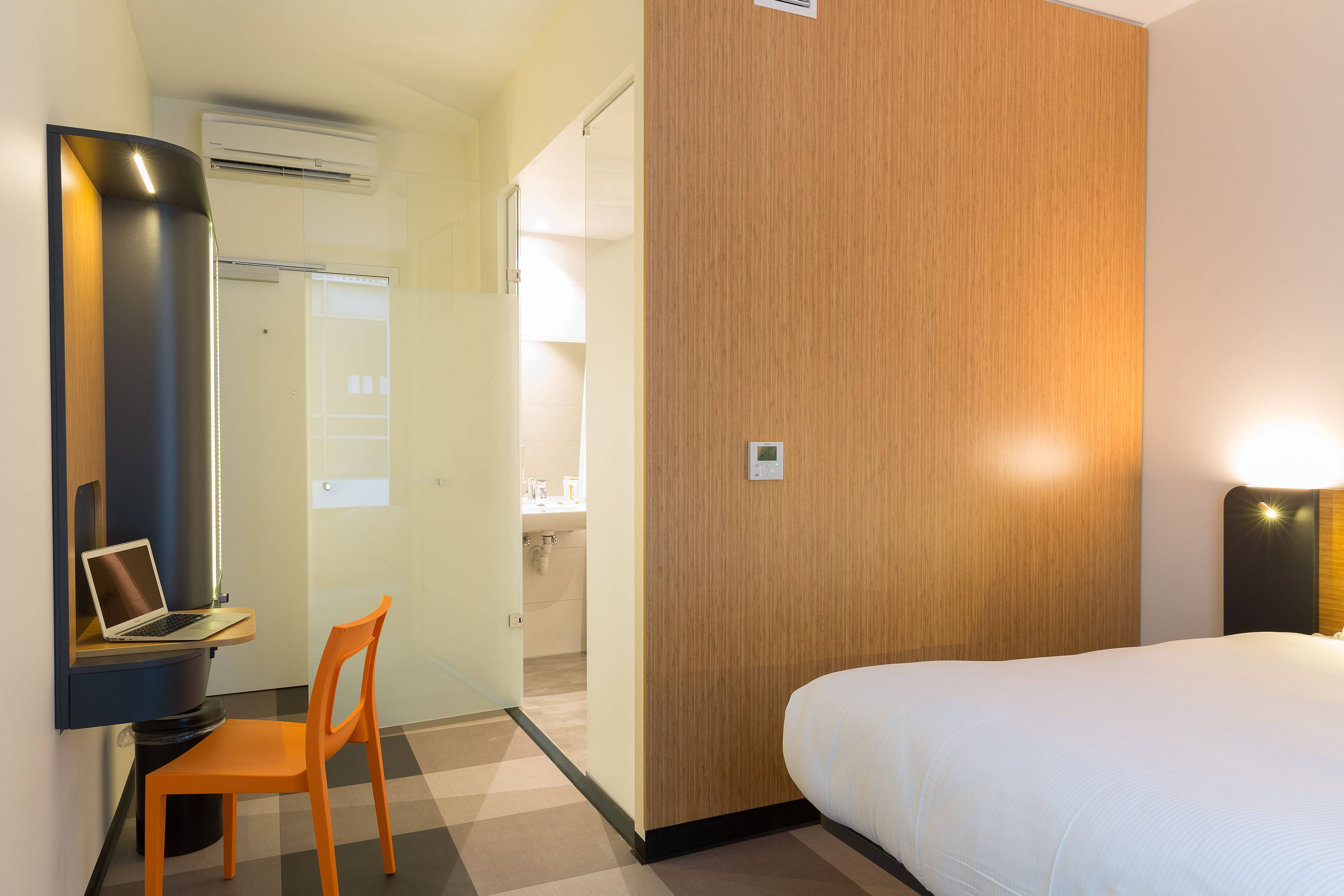 A hotel room with a bed, a desk with a laptop, an orange chair, a wooden accent wall, and a bathroom visible through an open door.