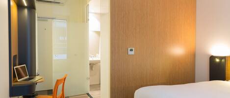 A hotel room with a bed, a desk with a laptop, an orange chair, a wooden accent wall, and a bathroom visible through an open door.