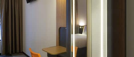 A hotel room with a large mirror, a wooden desk, an orange chair, a television mounted on the wall, and a black trash bin.